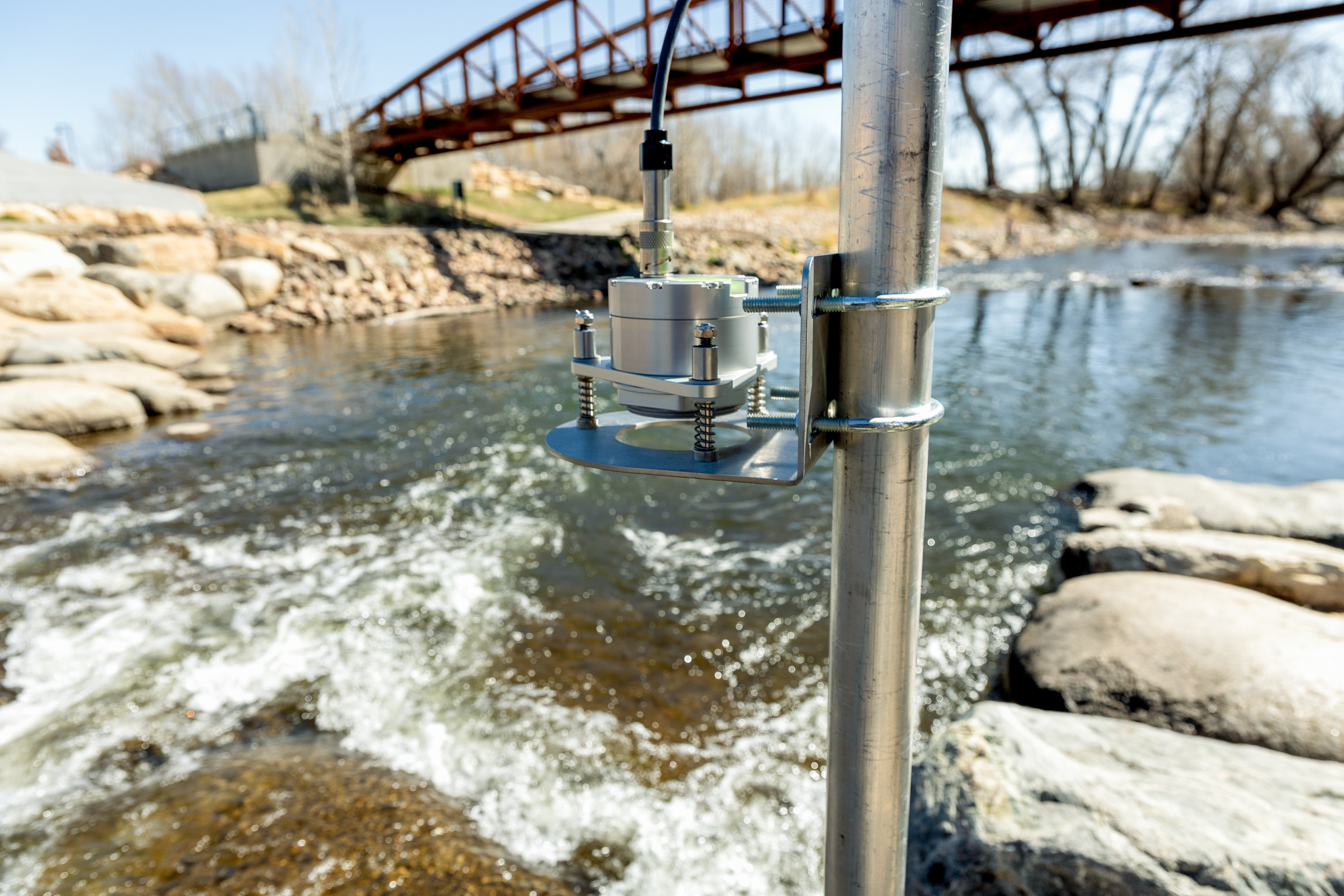 A water monitoring device next to a river bank with a bridge in the background.